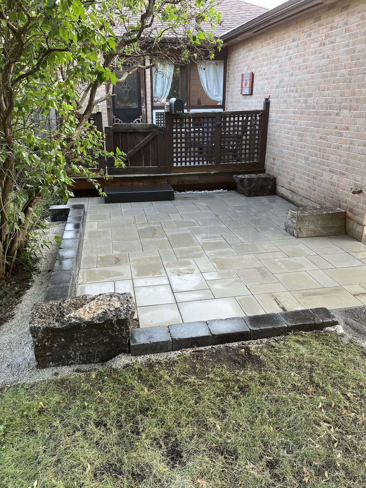 Wide view of the paver patio with multiple large rough-cut armour stone boulders stacked as natural seating at the back corner where the brick house meets a lattice fence