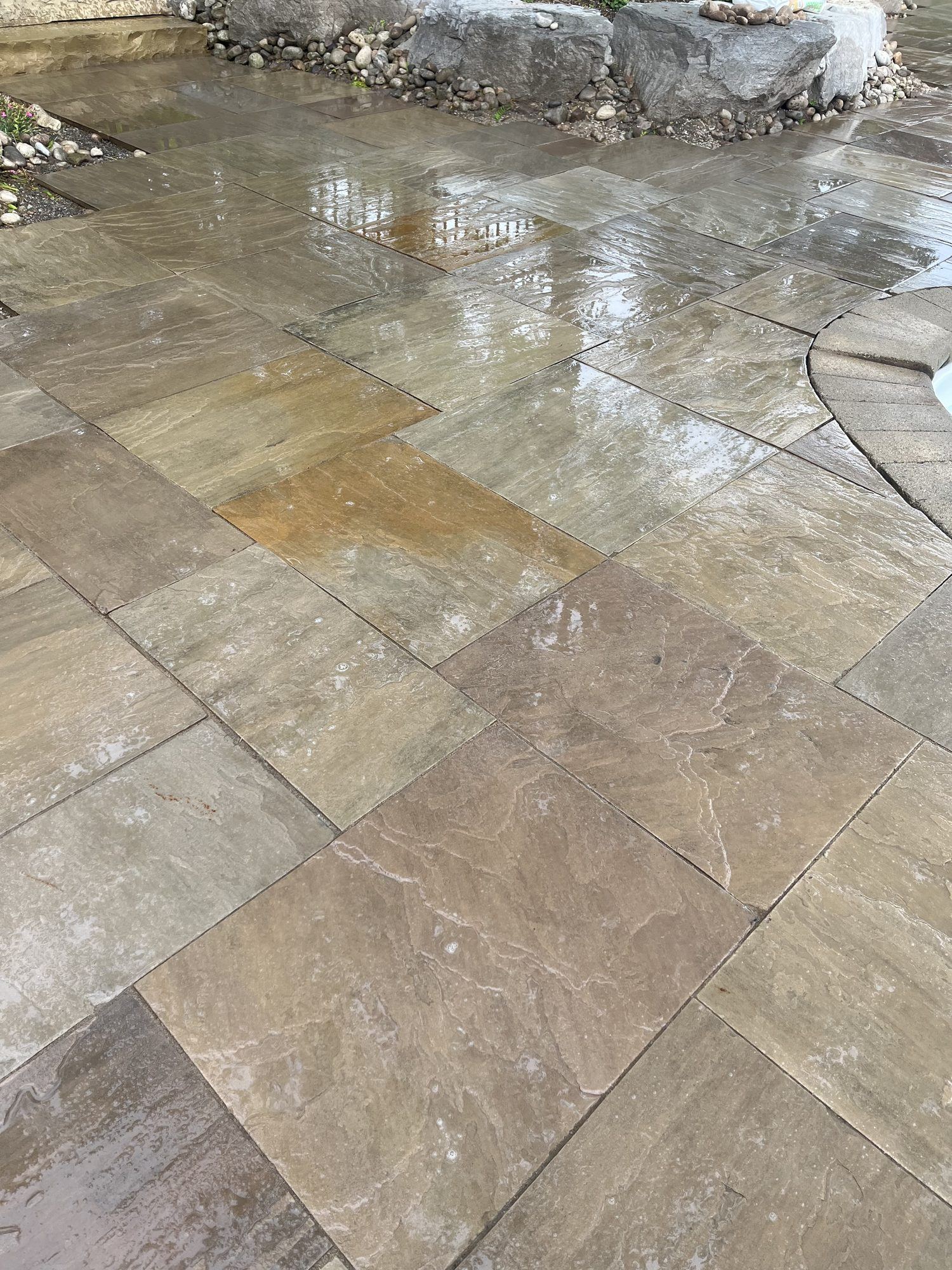 Wet natural sandstone pool deck laid in large rectangular slabs, curving around the edge of an in-ground pool with a river-rock border along the garden