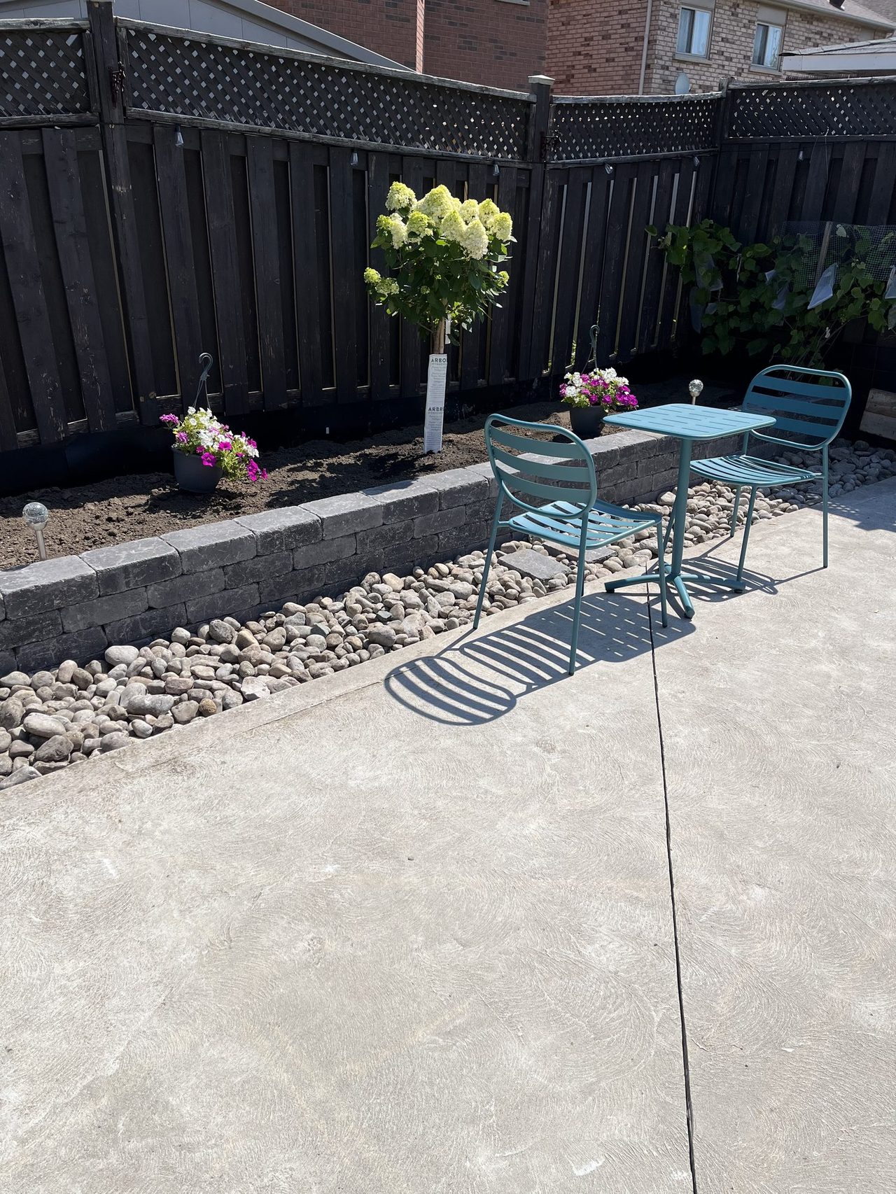 Charcoal split-face block raised planter with smooth slate-grey coping stones along a concrete patio, a river-rock border and a hydrangea tree with petunias in the bed