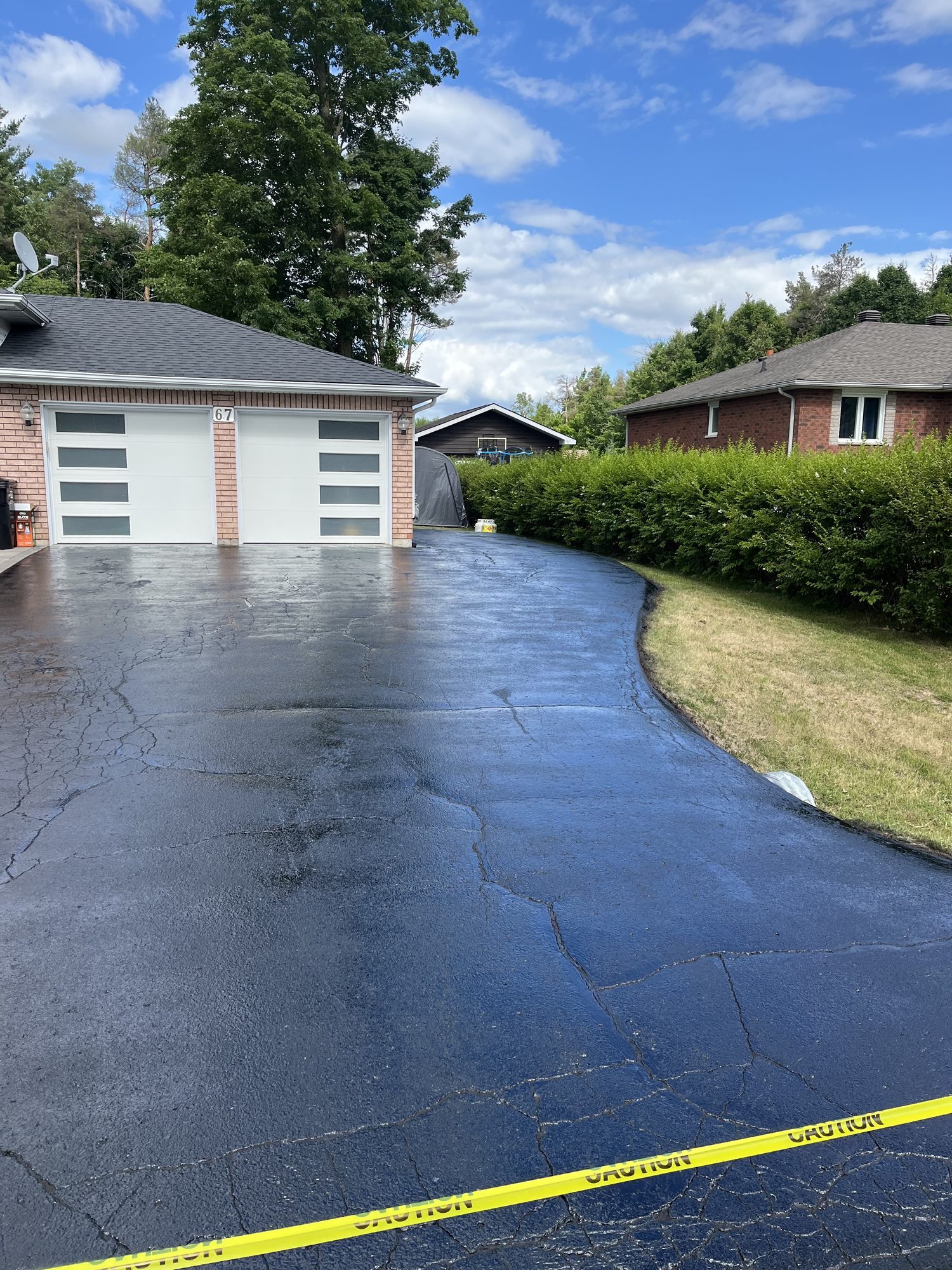 Freshly sealed black asphalt driveway in front of a two-car garage, glossy and still wet with yellow caution tape stretched across, deep cracks visible underneath the new coat