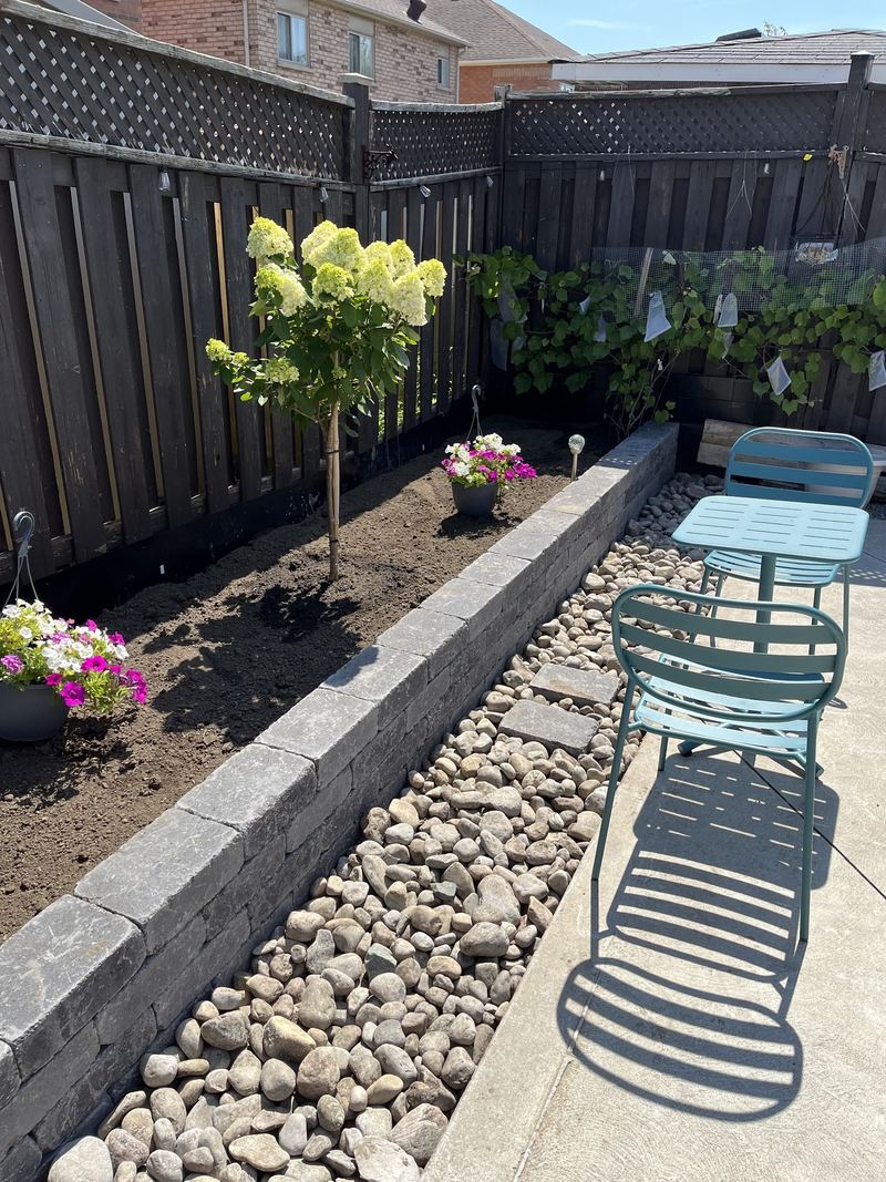 Raised planter and river-rock strip built in wall block along a backyard fence, planted with a young tree and flowers