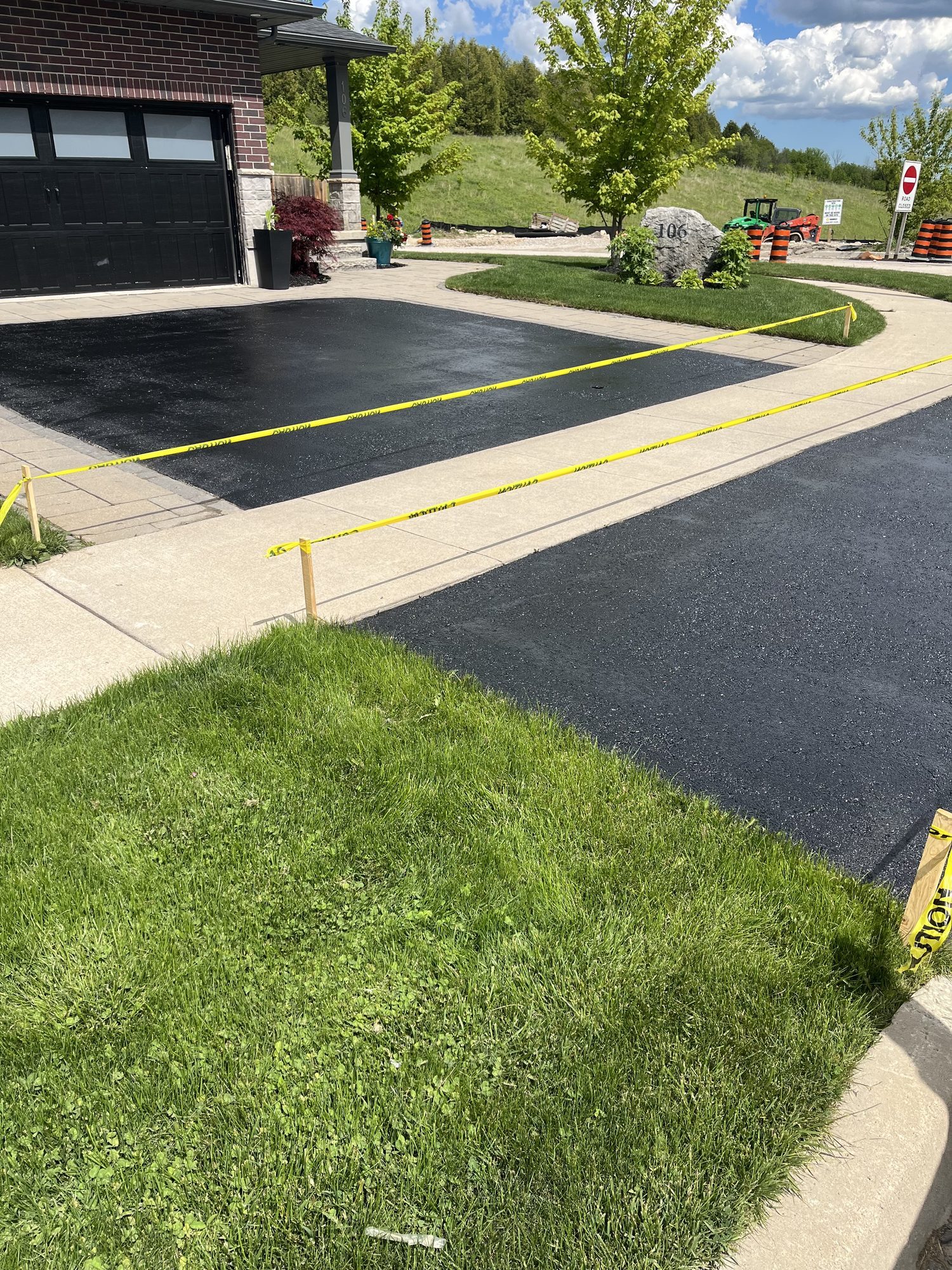 Newly sealed black asphalt driveway in front of a red brick house with a black garage door, bordered by light stone pavers and cordoned with caution tape on stakes