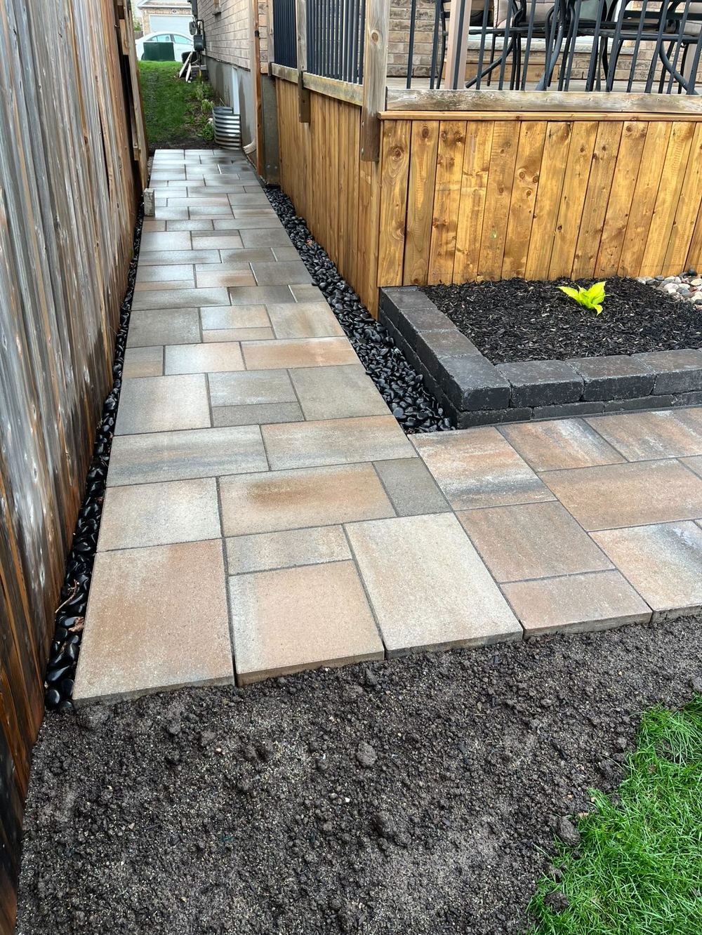 Side-yard slab walkway between a fence and a deck, with a low block-edged raised garden bed and fresh soil alongside