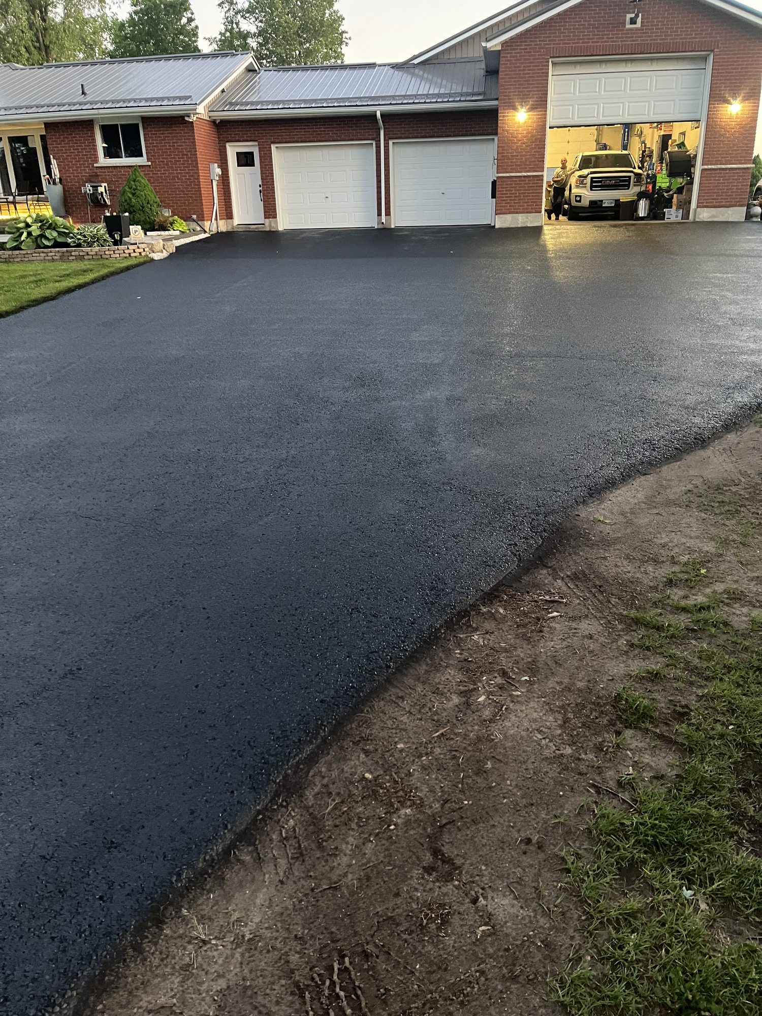 Wide freshly sealed charcoal-black asphalt driveway leading to a red brick house with three white garage doors, one open showing a white pickup truck, in soft evening light