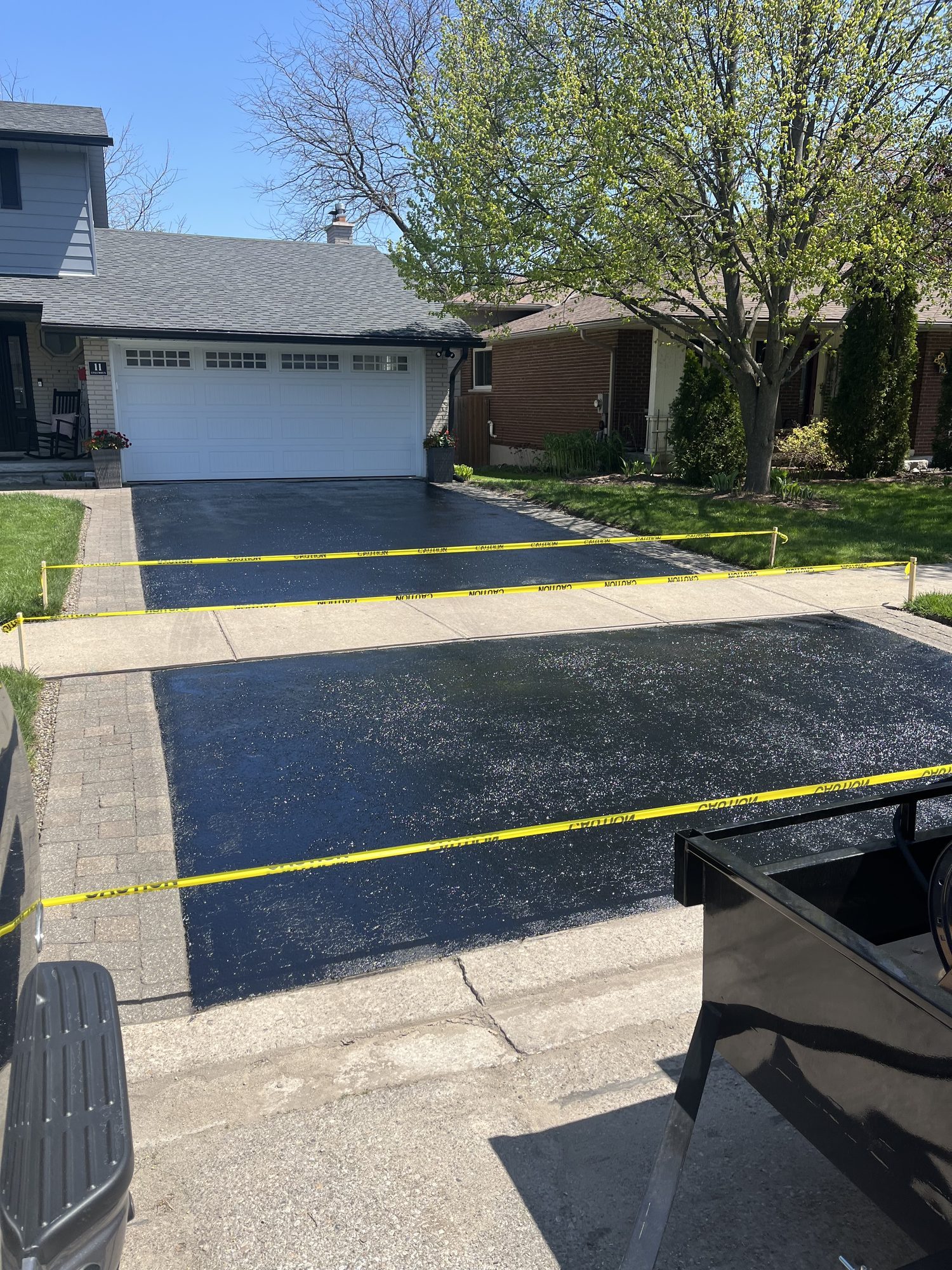 Freshly sealed jet-black asphalt driveway in two sections divided by a concrete sidewalk, cordoned off with yellow caution tape tied to wooden stakes across both bays