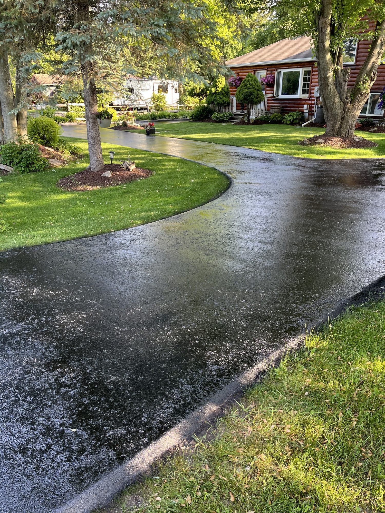 Freshly sealed black asphalt driveway curving in an S through a green lawn toward a brown log house with flower boxes under a bright summer sky