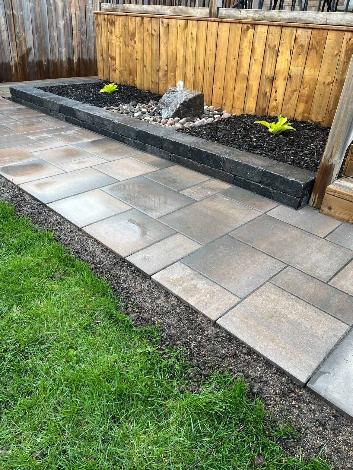 Paver walkway running beside a low charcoal block raised planter with a natural stone boulder water feature bubbling into river pebbles, hostas planted in the bed