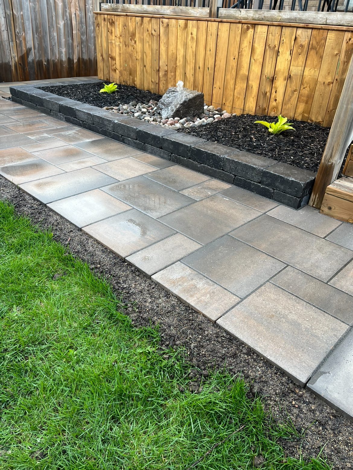 Wide view of the charcoal block raised planter and boulder water feature beside the paver walkway, backed by a wooden deck and fence