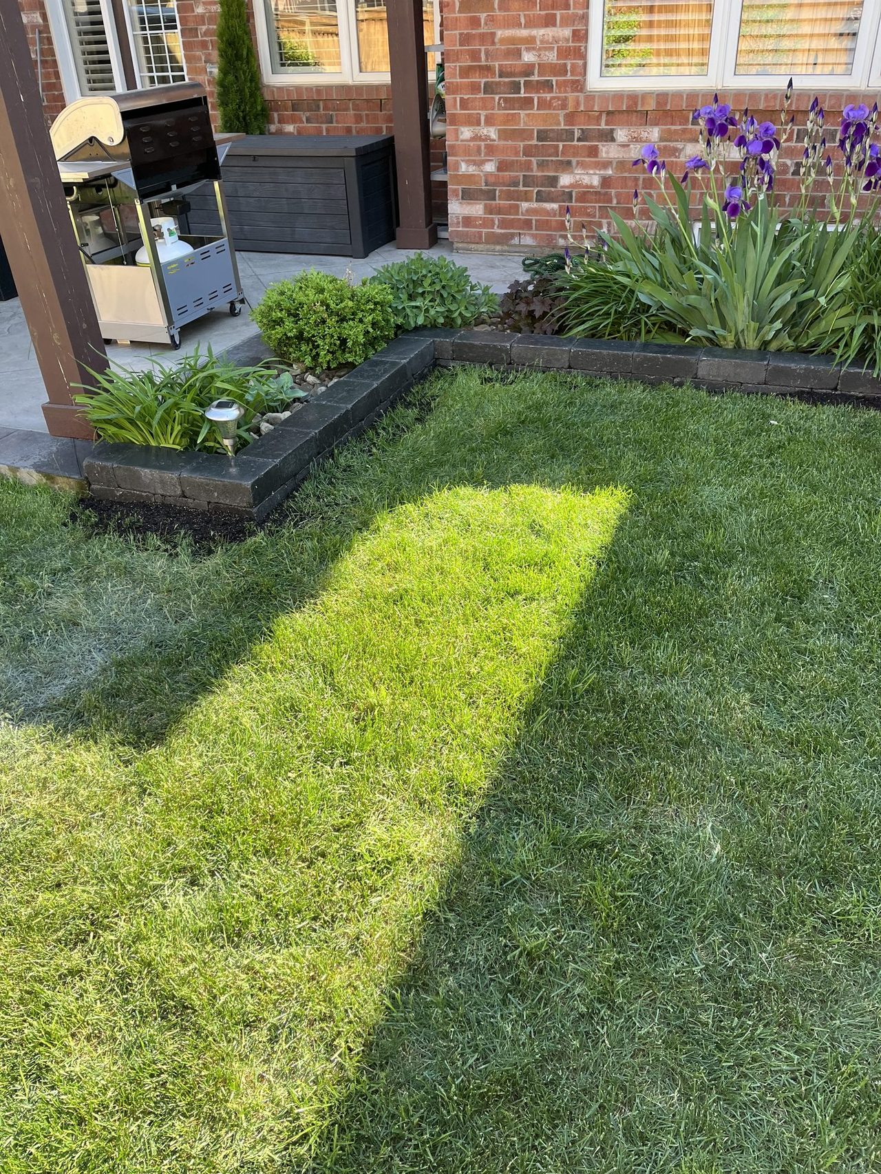 Two-course garden bed edging in charcoal and grey block forming an L-shaped corner around a planting bed with irises, beside a stone patio with a grill and pergola post