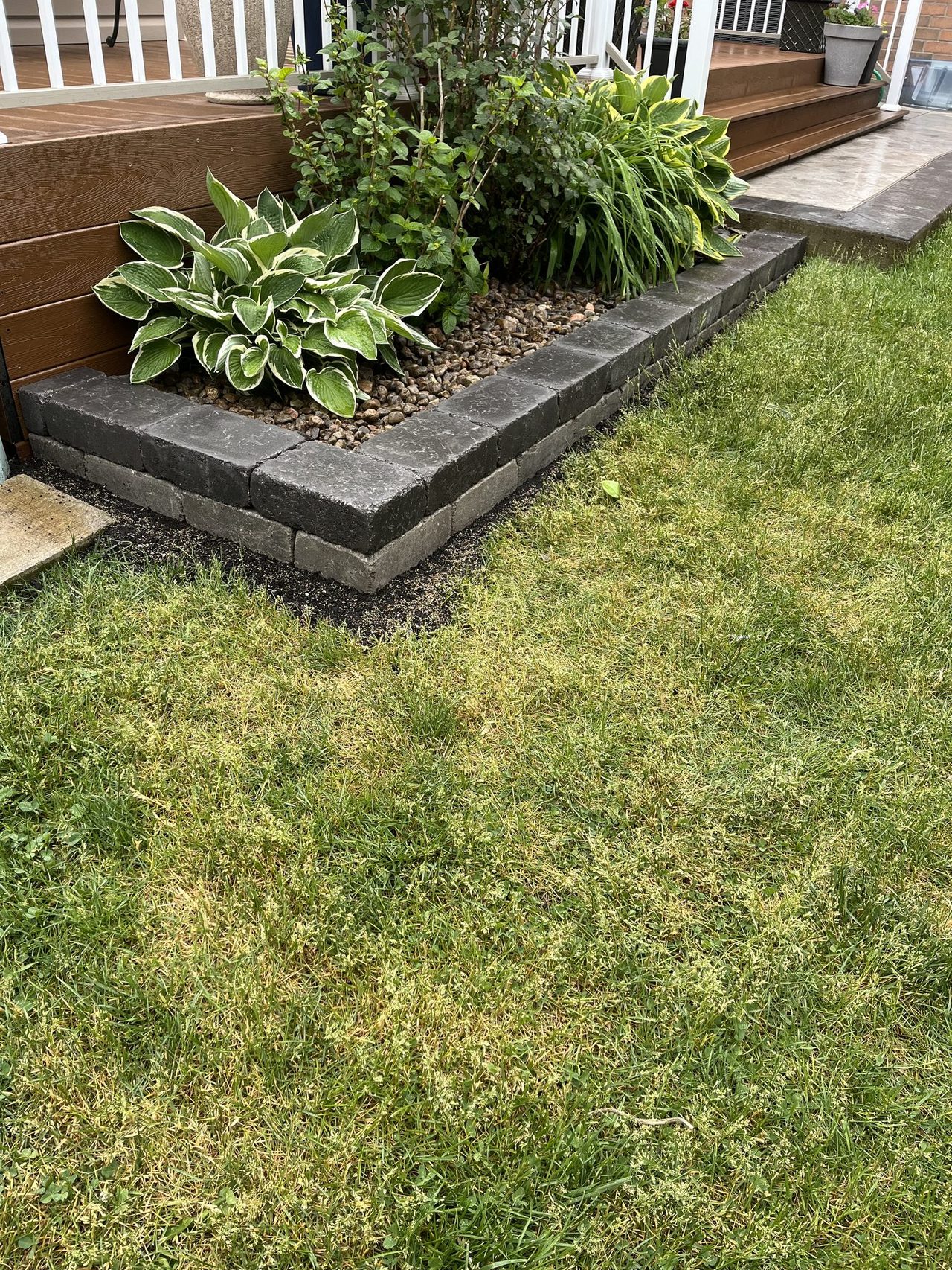 Two-course charcoal and grey block garden edging turning a 90-degree corner at the base of a white-railed wooden deck, the bed filled with river pebbles and hostas