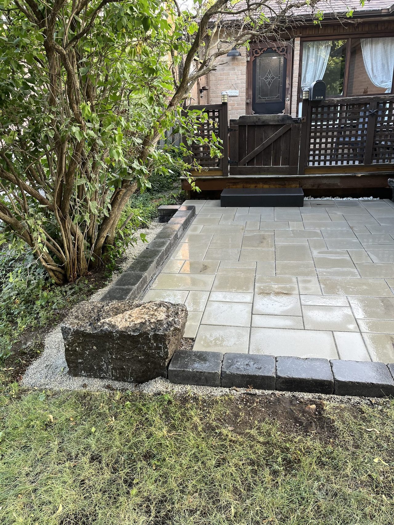 Light beige and grey paver patio with a charcoal cobble border course, anchored by a large rough-hewn natural armour stone boulder at the corner against a wooden deck