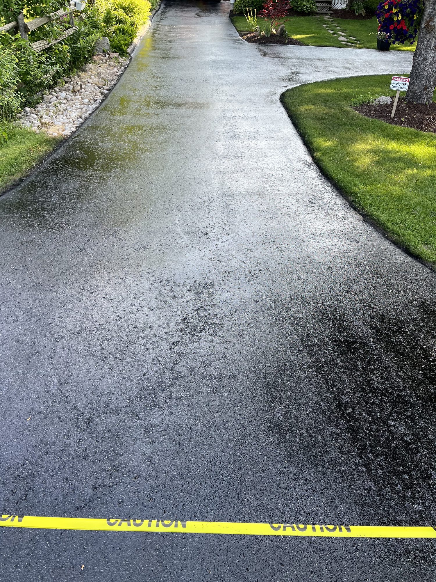 Freshly sealed black asphalt driveway cordoned with yellow caution tape, running past a wooden rail fence and a river-stone bed toward a lawn with a mature tree