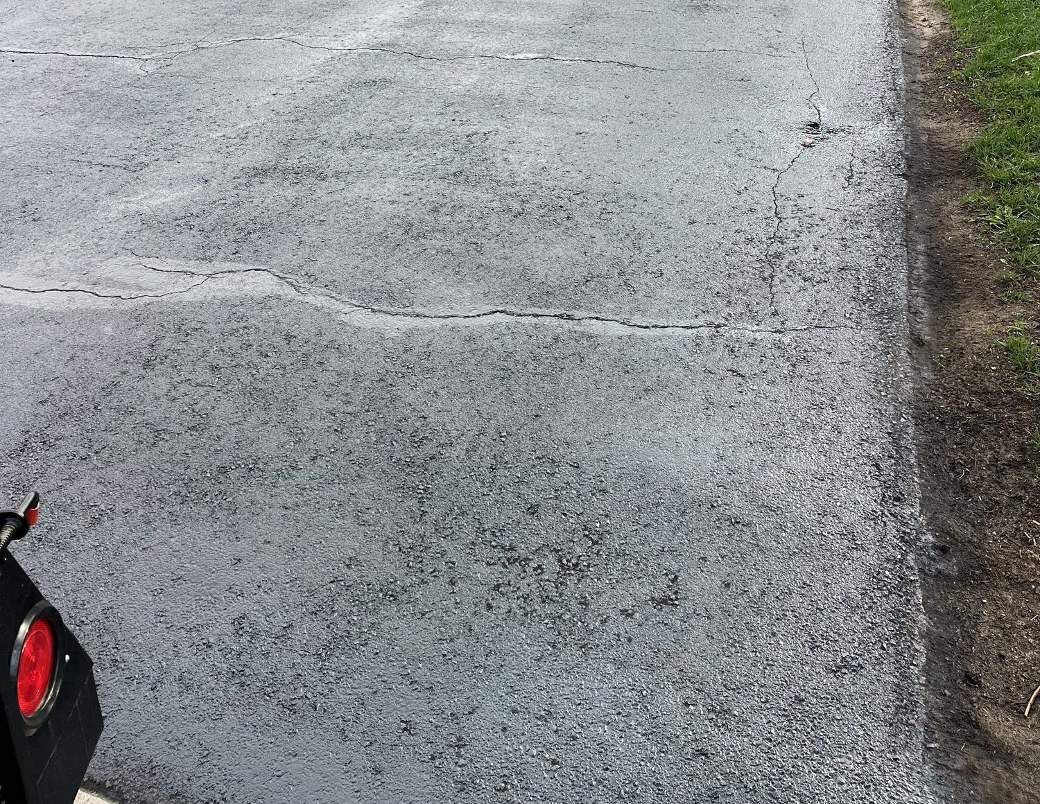 Before: faded grey asphalt driveway surface riddled with a network of wide cracks running across and along the pavement, bordered by grass and a soil edge